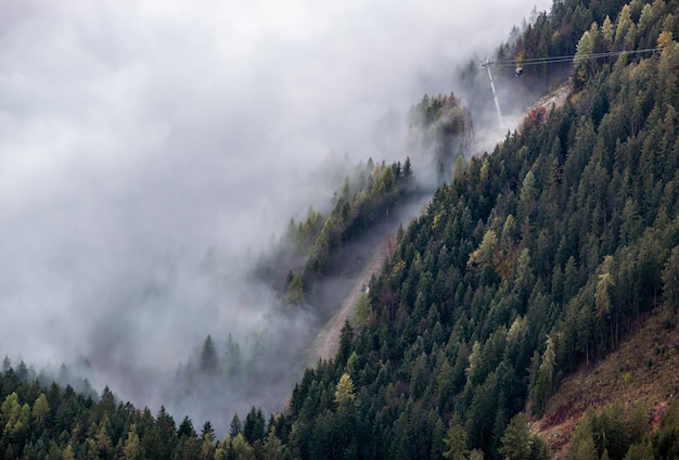 Gondole della funivia che si muovono attraverso la collina di montagna delle Alpi dalla nuvola nebbiosa alla cima Vista nebbiosa mattutina autunnale