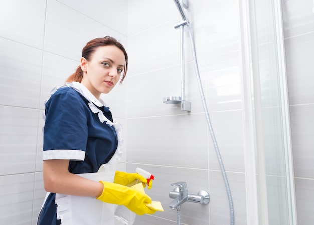 Governante che serve il bagno di un hotel in piedi sotto la doccia con indosso l'uniforme e il grembiule che guarda la telecamera