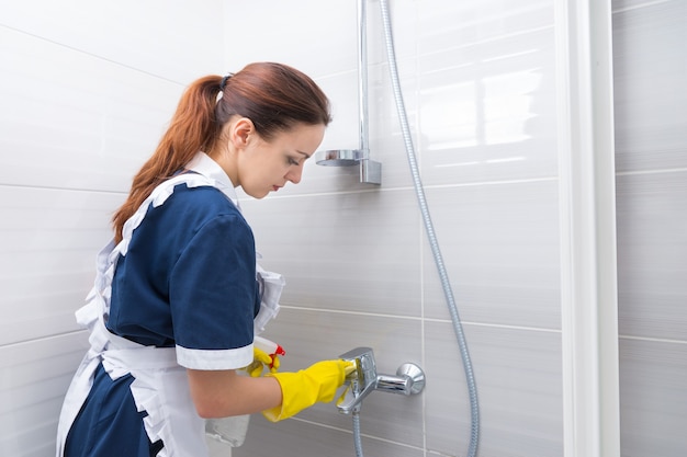 Governante in una casa di lusso che fa i lavori domestici in piedi sotto la doccia nella sua uniforme che pulisce il rubinetto