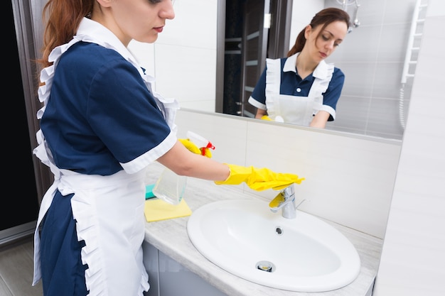 Governante in uniforme ordinata e grembiule che serve un bagno dell'hotel che lava e pulisce il lavabo e il rubinetto con un panno giallo, riflesso nello specchio