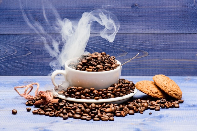 Grado di tostatura del grano. Menu delle bevande del caffè. Chicchi di caffè tostati freschi. Caffè per ispirazione e carica di energia. Tazza piena di caffè marrone chicchi tostati nuvole bianche di fumo blu sullo sfondo di legno.