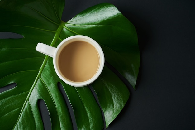 Grande derisione alla moda con caffè in una tazza bianca su sfondo nero con una foglia tropicale verde.