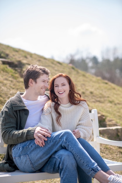 Grande fine settimana. Sorridente gioioso giovane che tocca la donna in abiti casual seduto su una panchina nella natura in una bella giornata