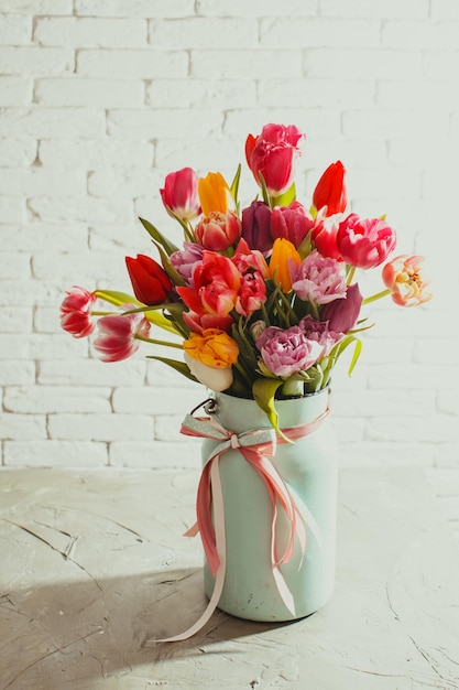 Grande mazzo di tulipani freschi in metallo smaltato può con nastro, in piedi sul tavolo bianco. Muro con trama di mattoni bianchi su sfondo, ombra di oggetti sul lato sinistro