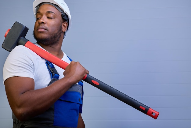 Grande ragazzo nero in un casco protettivo e abiti da lavoro guarda la mazza che giace sulla sua spalla potente lavoratore afroamericano in tuta su sfondo grigio