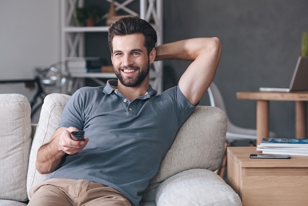 Grande spettacolo televisivo. Bel giovane allegro che tiene il telecomando e guarda la TV mentre è seduto sul divano di casa