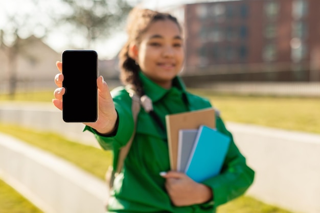 Grande studentessa di app educativa che mostra lo schermo vuoto del cellulare in piedi con cartelle di lavoro all'aperto