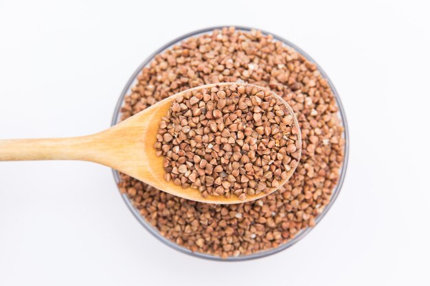 Grano del grano saraceno con il cucchiaio di legno in ciotola di vetro su fondo bianco. Isoalted su bianco