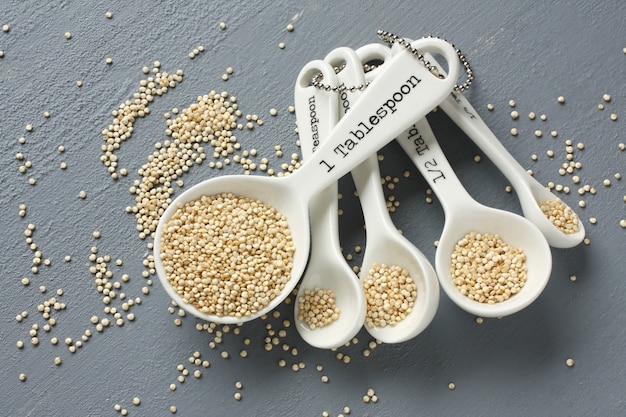 Grano della quinoa in porcellana che misura i cucchiai su fondo grigio