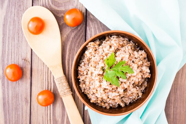 Grano saraceno cucinato in una ciotola con una foglia di prezzemolo, cucchiaio di legno e pomodori ciliegia su una tavola di legno, vista superiore