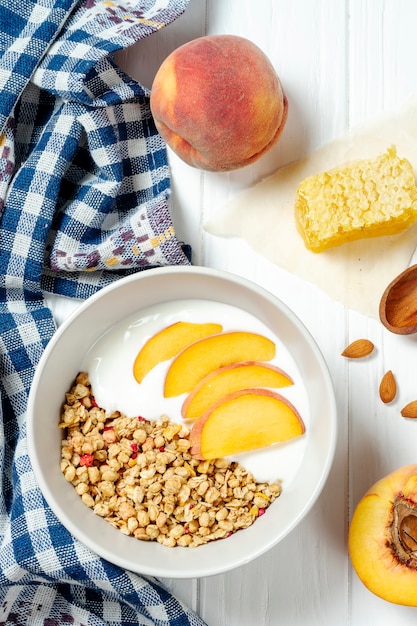 Granola di grano bianco con yogurt magro in una ciotola bianca in una composizione con un cucchiaio, favi e pesca su fondo di legno bianco.