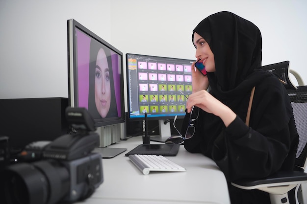 Graphic designer femminile musulmana che parla al telefono. Libero professionista che modifica le foto su due monitor. Ragazza hijab sul posto di lavoro