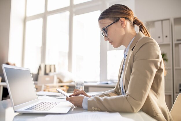 Grave concentrato giovane donna con gli occhiali seduto alla scrivania e controllare le note sullo smartphone mentre si lavora al progetto online