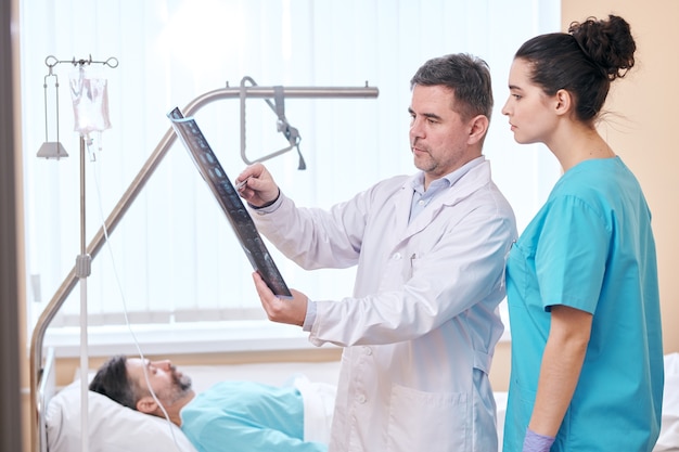 Grave medico maturo in camice da laboratorio che punta all'immagine a raggi x mentre discute i risultati con l'infermiera nella stanza dei pazienti
