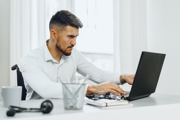 Grave uomo disabile concentrato in sedia a rotelle utilizzando il suo laptop per lavoro alla ricerca di un lavoro in internet