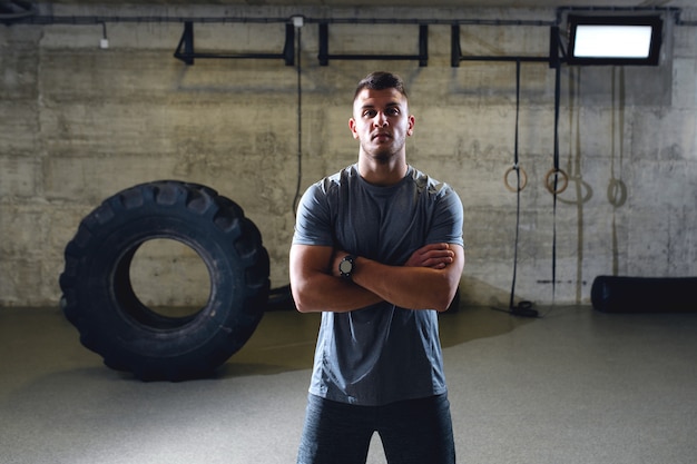 Grave uomo muscoloso in abiti sportivi in piedi con le braccia incrociate in palestra cross-fit.