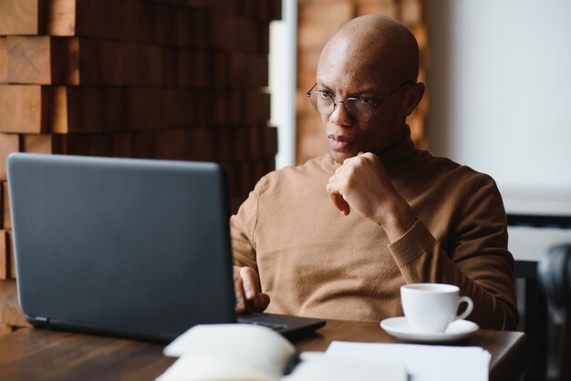 Gravi occhiali studente maschio nero prendere appunti mentre è seduto al tavolo con il computer portatile e lavorando al progetto in un accogliente caffè