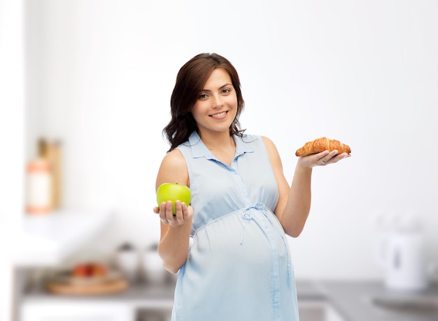 gravidanza, alimentazione sana, cibo spazzatura e concetto di persone - felice donna incinta che sceglie tra mela verde e croissant su sfondo cucina