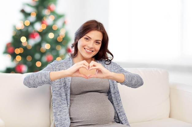 gravidanza, amore, vacanze, persone e concetto di aspettativa - felice donna incinta seduta sul divano e che fa il gesto del cuore sullo sfondo dell'albero di natale