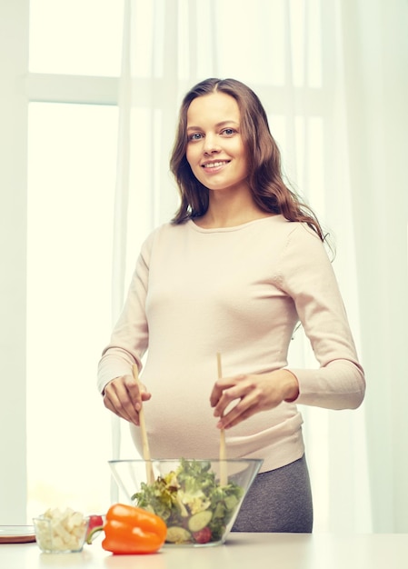 gravidanza, cucinare cibo, stile di vita sano, persone e concetto di aspettativa - donna incinta felice che mescola insalata di verdure in una ciotola a casa