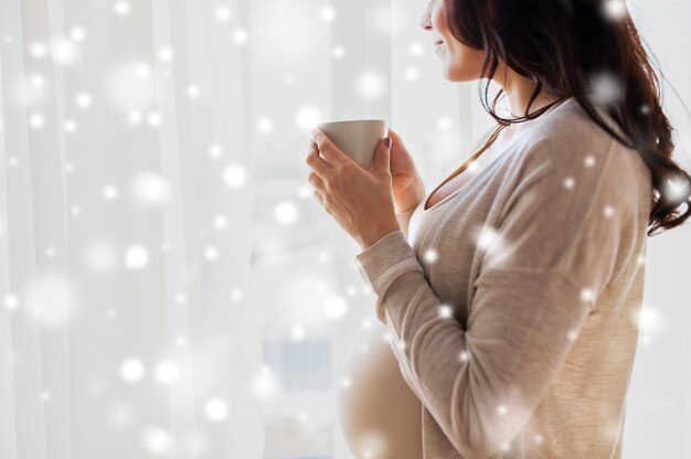 gravidanza, inverno, natale, persone e concetto di aspettativa - primo piano di una donna incinta felice con una tazza che beve tè guardando attraverso la finestra a casa sopra la neve