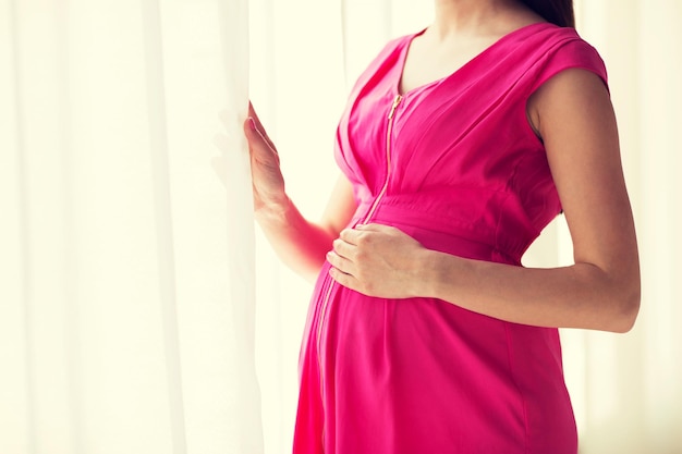 gravidanza, maternità, persone e concetto di aspettativa - primo piano di una donna incinta che guarda attraverso la finestra a casa