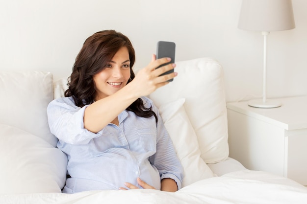 Gravidanza, maternità, tecnologia, persone e concetto di aspettativa - donna incinta felice con smartphone che si fa selfie a letto a casa