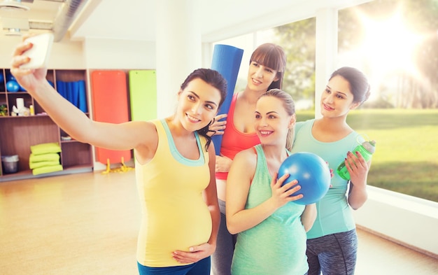 gravidanza, sport, fitness, persone e concetto di stile di vita sano - gruppo di donne incinte felici con cose sportive che prendono selfie con lo smartphone in palestra su sfondo naturale della finestra