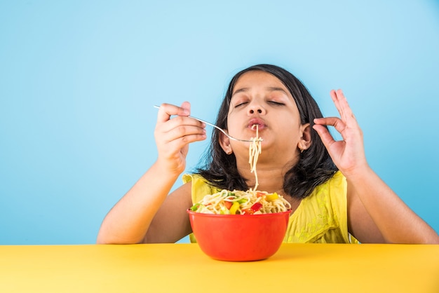Graziosa bambina indiana o asiatica che mangia gustosissime tagliatelle cinesi con forchetta o bacchette, isolate su sfondo colorato