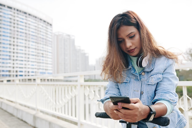 Graziosa giovane donna in piedi su uno scooter e controlla i messaggi di testo e le notifiche sul suo telefono