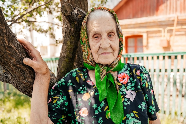 Grazioso ritratto di una donna anziana vestita con accappatoio e sciarpa in posa per una foto vicino a un vecchio albero nel suo cortile