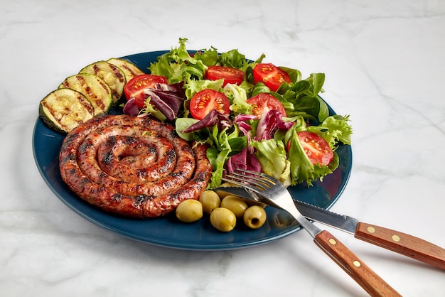 Grigliata di salsiccia a spirale con pomodori a fette, zucchine e lattuga su un piatto blu su un tavolo luminoso