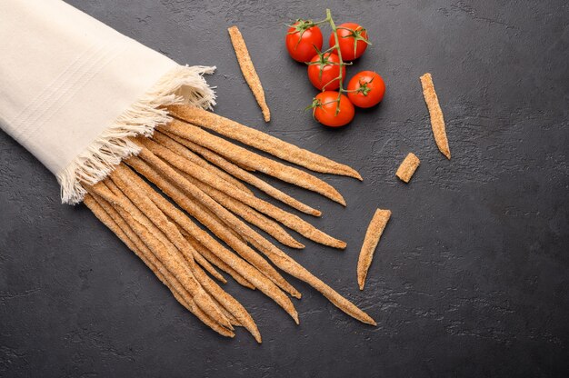 Grissini di pane di segale tradizionale italiano e pomodorini sul tovagliolo di lino su sfondo scuro.