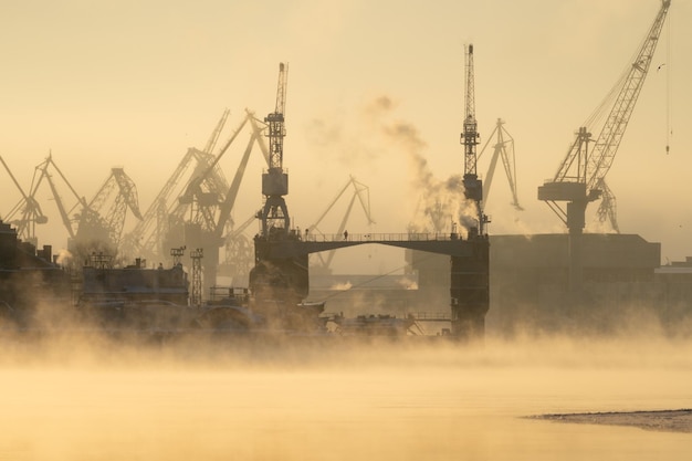 Gru del cantiere navale nella gelida giornata invernale a vapore sulla superficie liscia del fiume al tramonto