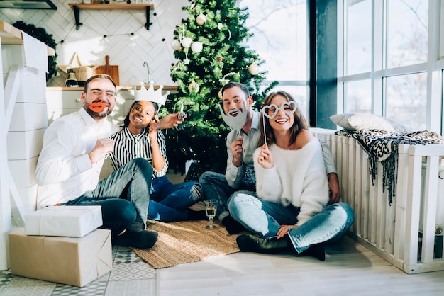 Gruppo allegro di amici che guarda l'obbiettivo vicino all'albero di abete in soggiorno decorato per la vigilia di Natale