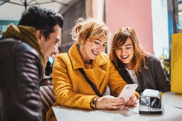 Gruppo di adulti allegri seduti al bar a guardare contenuti sullo schermo dello smartphone - Amico adulto che sorride navigando sui social network seduto al bar all'aperto