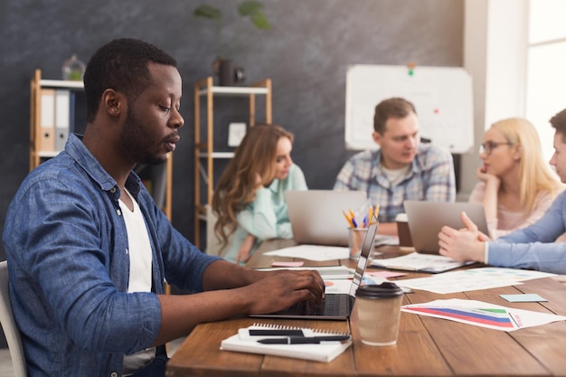 Gruppo di affari che parla dei risultati del loro lavoro. Gruppo di uomini d'affari allegri che discutono di una nuova strategia di marketing, giovane uomo di colore che digita sul computer portatile, copia spazio