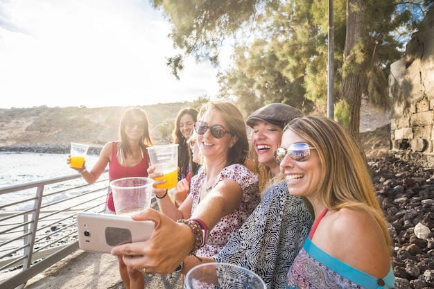 Gruppo di amiche che si fanno selfie sullo smartphone mentre tengono in mano un bicchiere di succo di mare