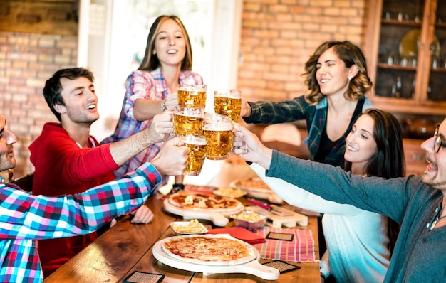 Gruppo di amici che bevono birra e mangiano pizza al bar ristorante - Concetto di amicizia con i giovani che si divertono insieme al risto pub pizzeria durante l'happy hour - Focus sui bicchieri da pinta - Filtro caldo