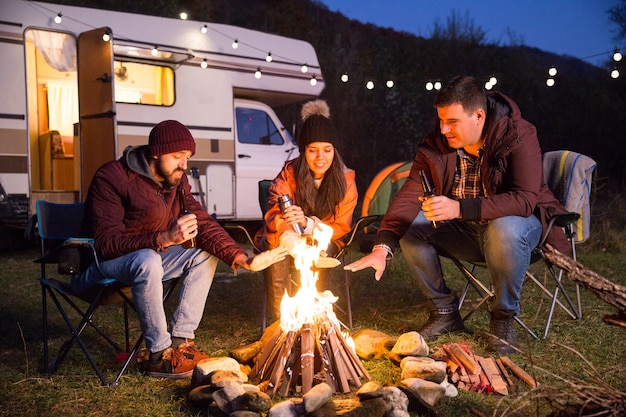 Gruppo di amici che si godono l'aria fresca delle montagne intorno al fuoco da campo. Camper retrò con lampadine.