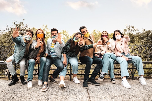 Gruppo di amici di generazione millenaria che indossano una maschera facciale facendo ok e v come segni di vittoria seduti su una panchina nel parco