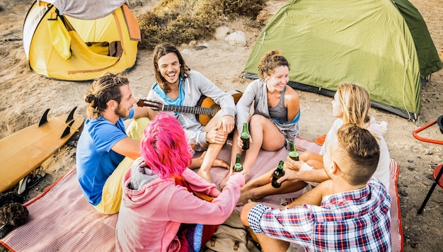 Gruppo di amici divertendosi insieme alla festa in campeggio sulla spiaggia - concetto di viaggio di amicizia felice con giovani travalers suonare la chitarra e bere birra in bottiglia al surf camp estivo