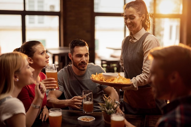 Gruppo di amici felici che bevono birra in una taverna mentre la cameriera serve loro patatine al nacho