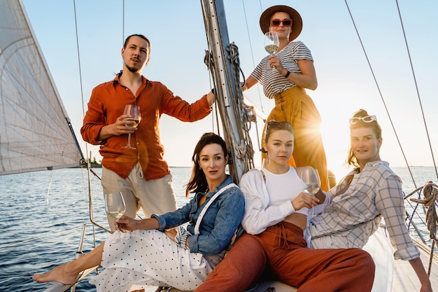 Gruppo di amici felici che bevono vino e si rilassano sulla barca a vela durante la navigazione in mare