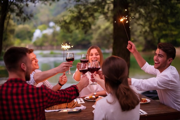 gruppo di amici felici che celebrano le vacanze usando irrigatori e bevendo vino rosso mentre fanno un picnic con cena francese all'aperto vicino al fiume in una bella serata estiva nella natura