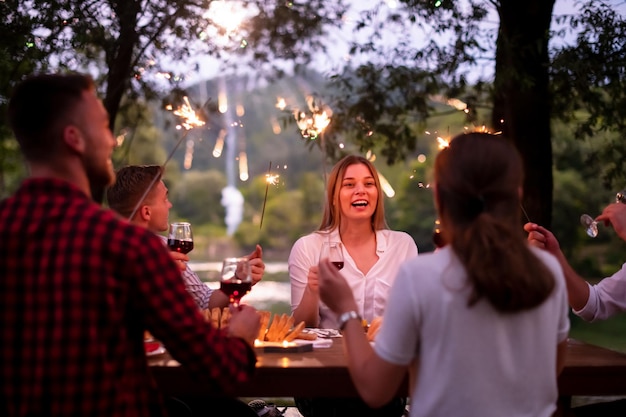 gruppo di amici felici che celebrano le vacanze usando irrigatori e bevendo vino rosso mentre fanno un picnic con cena francese all'aperto vicino al fiume in una bella serata estiva nella natura