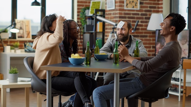 Gruppo di amici felici che giocano a giochi da tavolo con le carte, divertendosi insieme a bottiglie di birra e snack sul tavolo. I giovani si sentono allegri con il gameplay per l'intrattenimento. Scatto da treppiede.