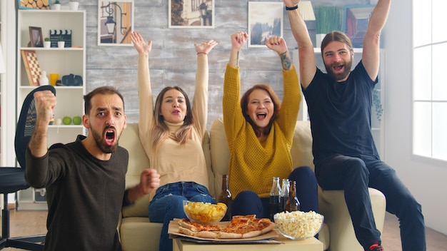 Gruppo di amici felici dopo che la loro squadra di calcio preferita ha vinto il campionato. Amici seduti sul divano a mangiare pizza e patatine.