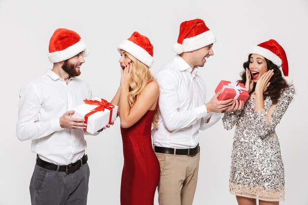 Gruppo di amici felici vestiti con cappelli rossi in piedi isolato su bianco, celebrando il nuovo anno, scambiando con i regali