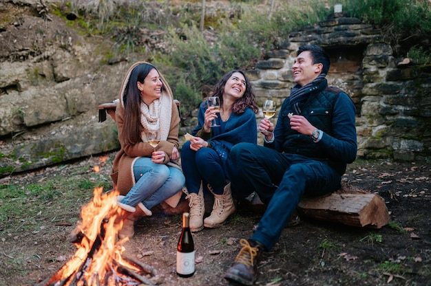 Gruppo di amici seduti davanti a un falò bevendo un bicchiere di vino mentre parlano e ridono. Insieme, concetto di amicizia.